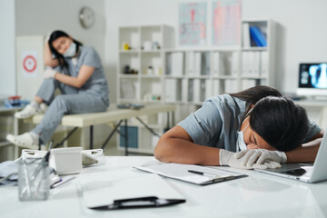 Obraz premium Young tired mixed-race female clinician in uniform and protective workwear napping on desk
