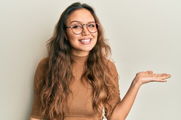 Young hispanic girl wearing casual clothes and glasses smiling cheerful presenting and pointing with palm of hand looking at the camera.