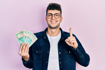 Young hispanic man holding united arab emirates dirham banknotes smiling with an idea or question pointing finger with happy face, number one