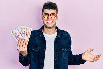 Young hispanic man holding saudi arabia riyal banknotes celebrating achievement with happy smile and winner expression with raised hand