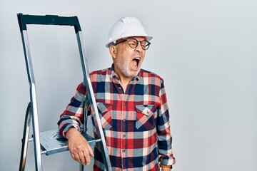 Handsome mature handyman close to construction stairs wearing hardhat angry and mad screaming frustrated and furious, shouting with anger. rage and aggressive concept.