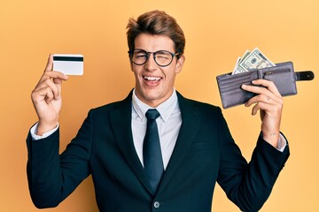 Handsome caucasian business man holding credit card and wallet with dollars winking looking at the...