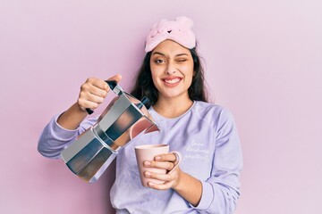 Beautiful middle eastern woman wearing sleep mask and pyjama drinking coffee winking looking at the camera with sexy expression, cheerful and happy face.