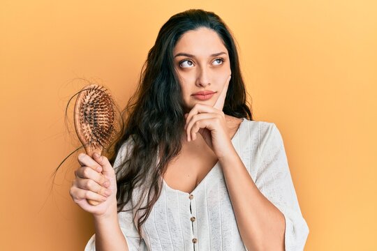 Beautiful middle eastern woman holding comb loosing hair serious face thinking about question with hand on chin, thoughtful about confusing idea