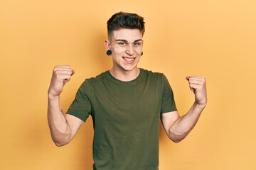 Young caucasian boy with ears dilation wearing casual green t shirt screaming proud, celebrating victory and success very excited with raised arms