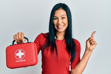 Beautiful hispanic woman holding first aid kit smiling happy pointing with hand and finger to the...