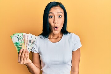 Beautiful hispanic woman holding russian ruble banknotes scared and amazed with open mouth for surprise, disbelief face