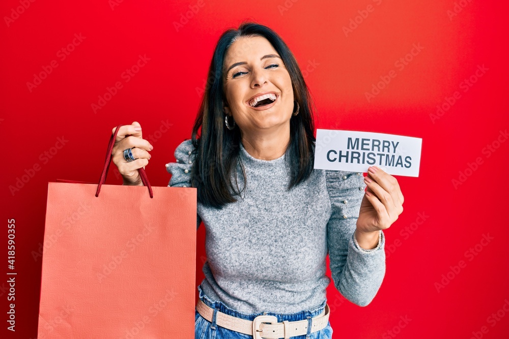 Poster middle age brunette woman holding merry christmas message and shopping bag smiling and laughing hard