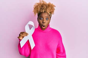 Young african woman with afro hair holding pink cancer ribbon scared and amazed with open mouth for...