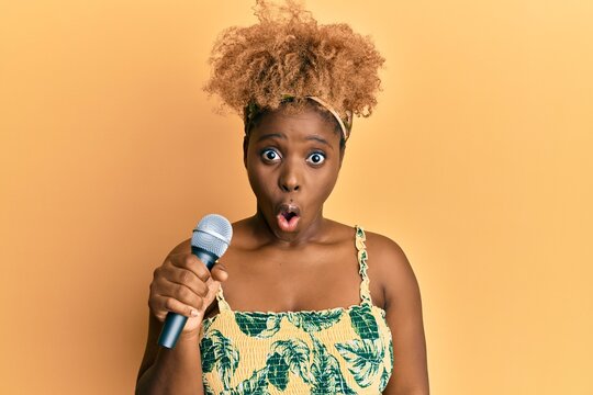 Young african woman with afro hair singing song using microphone scared and amazed with open mouth for surprise, disbelief face