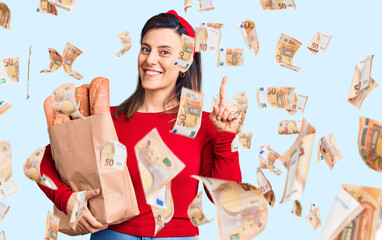 Young beautiful woman holding paper bag with bread surprised with an idea or question pointing finger with happy face, number one