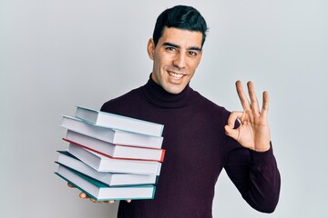 Handsome hispanic man holding a pile of books doing ok sign with fingers, smiling friendly gesturing excellent symbol