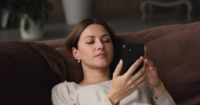 Relaxed happy young woman lying on cozy sofa, watching funny photo video content in social network, laughing enjoying pleasant distant communication in social network using mobile application.