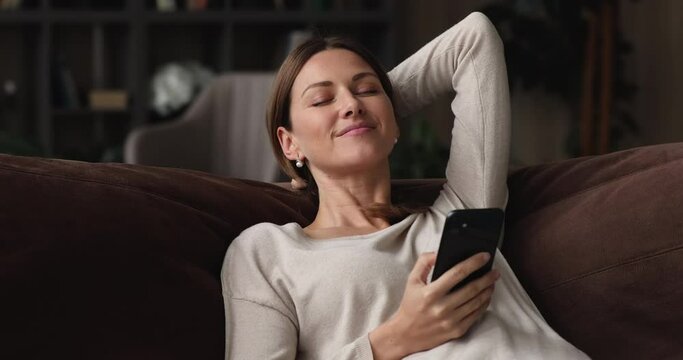 Happy attractive young 30s woman holding smartphone in hands, resting on comfortable sofa, enjoying stress free leisure moment alone in living room using mobile apps, daydreaming or napping indoors.