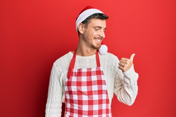 Handsome caucasian man wearing baker apron and christmas hat pointing thumb up to the side smiling...