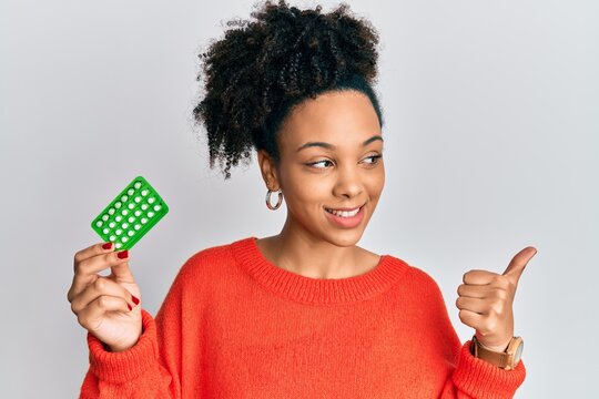 Young african american girl holding birth control pills pointing thumb up to the side smiling happy with open mouth