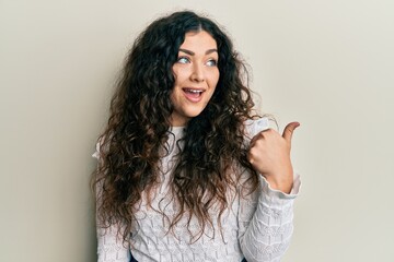 Fototapeta premium Young brunette woman with curly hair wearing casual clothes pointing thumb up to the side smiling happy with open mouth