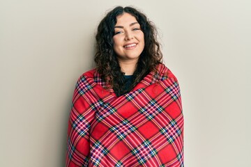 Fototapeta premium Young brunette woman with curly hair wrapped in a red warm red blanket smiling with a happy and cool smile on face. showing teeth.