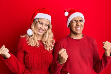 Young interracial couple wearing christmas hat very happy and excited doing winner gesture with arms raised, smiling and screaming for success. celebration concept.