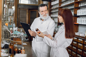 Confident female pharmacist talking with her senior male colleague about side effects of a medicine or pharmaceutical product for sale, working in old ancient pharmacy.