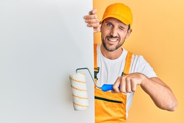 Young handsome man holding roller painter over white banner winking looking at the camera with sexy...