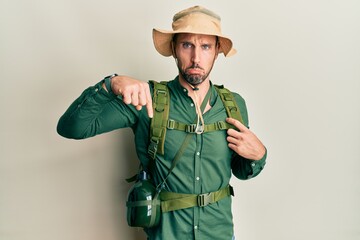 Handsome man with beard wearing explorer hat and backpack pointing down looking sad and upset, indicating direction with fingers, unhappy and depressed.