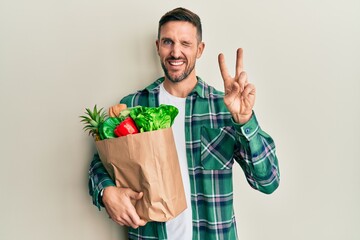 Handsome man with beard holding paper bag with groceries smiling with happy face winking at the...