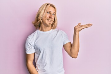 Caucasian young man with long hair wearing casual white t shirt smiling cheerful presenting and pointing with palm of hand looking at the camera.
