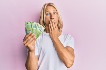 Caucasian young man with long hair holding 1000 chilean pesos covering mouth with hand, shocked and afraid for mistake. surprised expression