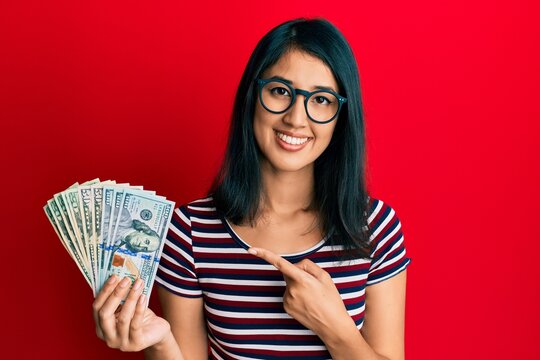 Beautiful asian young woman holding dollars smiling happy pointing with hand and finger