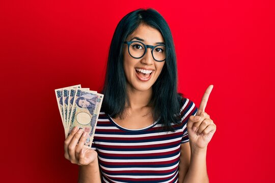 Beautiful asian young woman holding 5000 japanese yen banknotes smiling with an idea or question pointing finger with happy face, number one