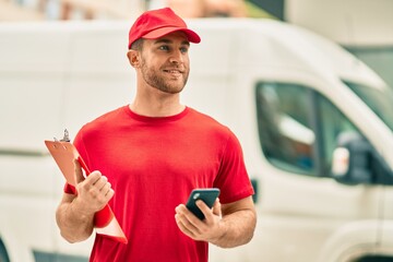 Young caucasian deliveryman using smartphone and holding clipboard at the city.
