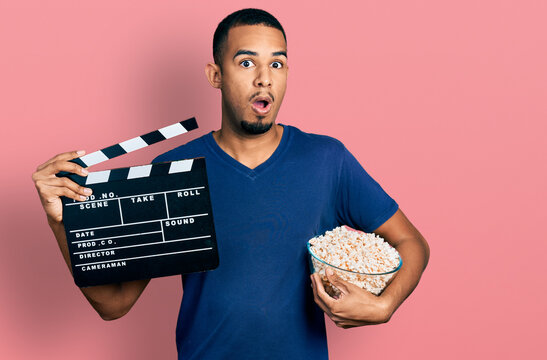 Young African American Man Eating Popcorn Holding Film Clapboard Afraid And Shocked With Surprise And Amazed Expression, Fear And Excited Face.