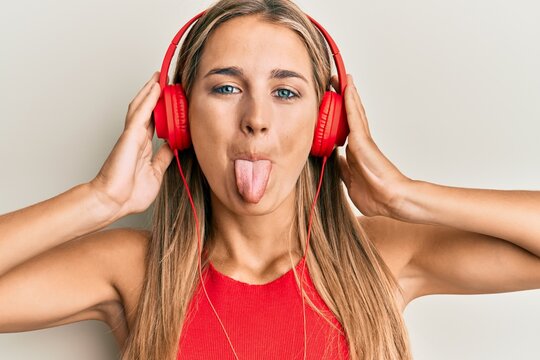 Young blonde woman listening to music using headphones sticking tongue out happy with funny expression.
