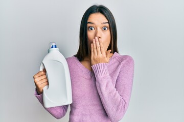 Young hispanic girl holding detergent bottle covering mouth with hand, shocked and afraid for...