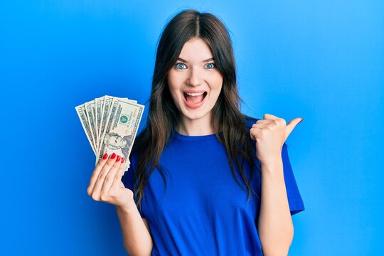 Young beautiful caucasian girl holding 20 dollars banknote pointing thumb up to the side smiling happy with open mouth