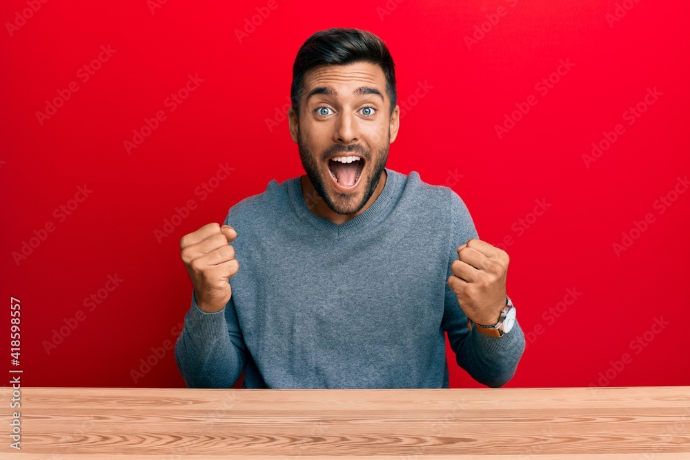 Wall mural Handsome hispanic man wearing casual style sitting on the table celebrating surprised and amazed for success with arms raised and open eyes. winner concept. - Wall murals
