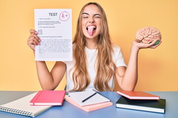 Young beautiful blonde woman holding brain and passed test sticking tongue out happy with funny...