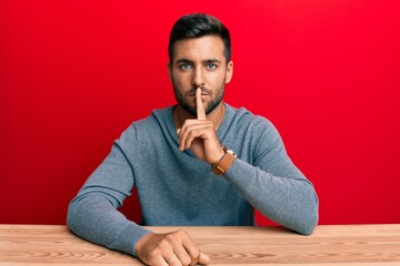 Handsome hispanic man wearing casual style sitting on the table asking to be quiet with finger on lips. silence and secret concept.