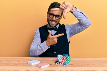 Handsome hispanic croupier man sitting on the table with poker chips and cards smiling making frame with hands and fingers with happy face. creativity and photography concept.