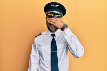 Handsome hispanic man wearing airplane pilot uniform smiling and laughing with hand on face covering eyes for surprise. blind concept.