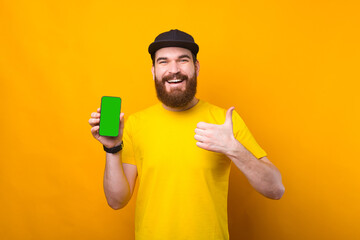 Portrait of cheerful young bearded man showing green screen on smartphone and showing thumb up