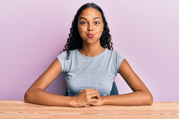 Young african american girl wearing casual clothes sitting on the table puffing cheeks with funny face. mouth inflated with air, crazy expression.