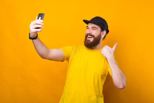 Photo of smiling man with beard over yellow background taking selfie and showing thumb up