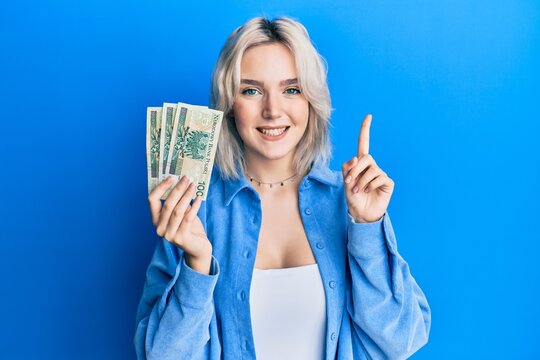Young blonde girl holding 50 polish zloty banknotes smiling with an idea or question pointing finger with happy face, number one