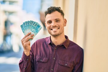 Young hispanic man smiling happy holding brazilian real banknotes at the city.
