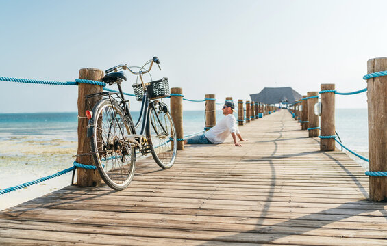 Old bicycle on wooden sea pier and a happy smiling man dressed in light summer clothes sitting and enjoying time on background. Careless vacation in tropical countries concept image. Zanzibar, Tanzani