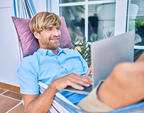 Middle age handsome man at the terrace of his house relaxing lying on a hammock working with laptop