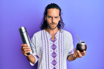 Young handsome man with long hair drinking mate infusion skeptic and nervous, frowning upset...