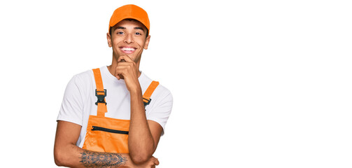 Young handsome african american man wearing handyman uniform looking confident at the camera with smile with crossed arms and hand raised on chin. thinking positive.